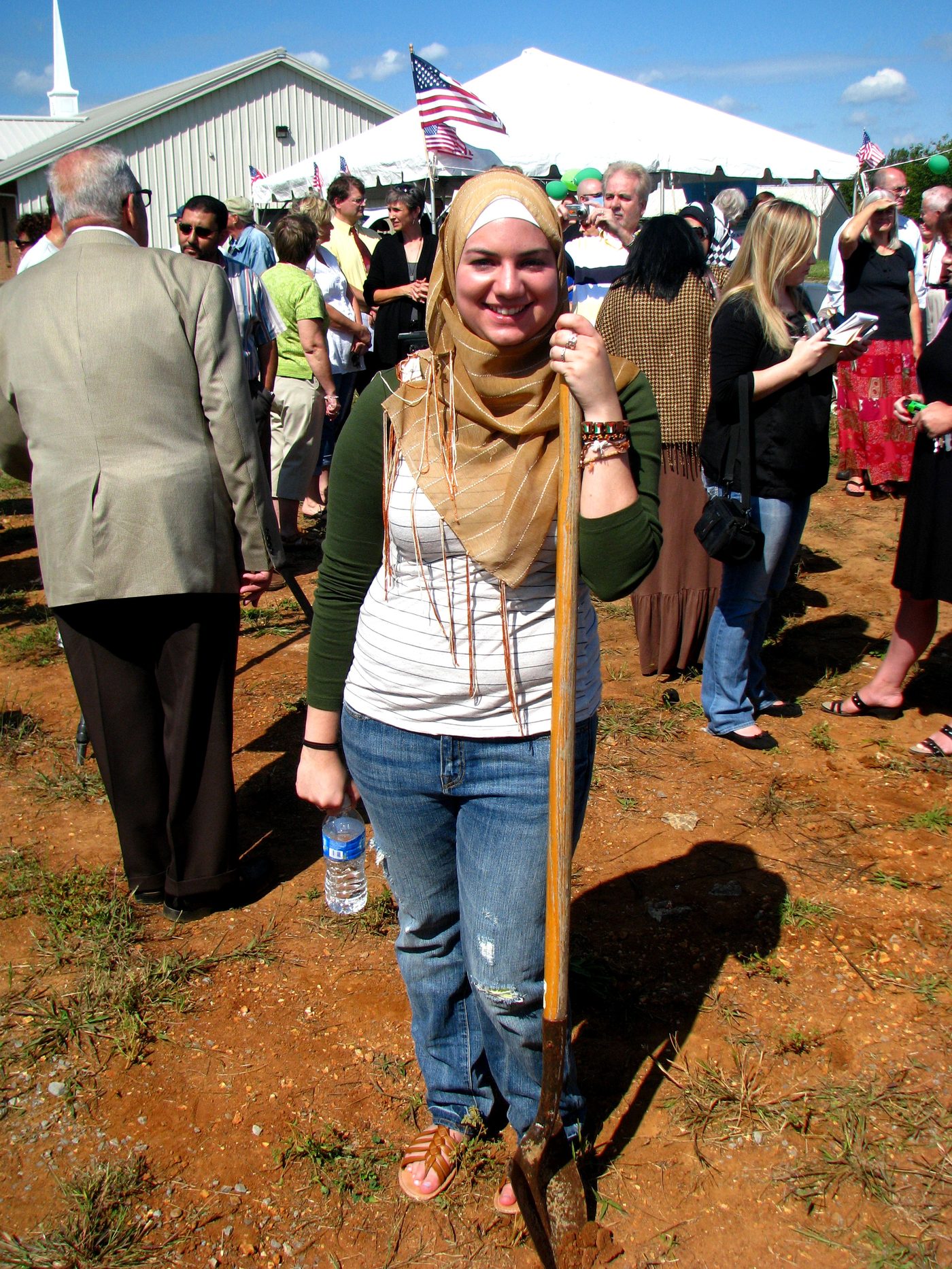 A young ICM community member smiling and holding a shovel at the 2010 groundbreaking ceremony, with a celebration tent and American flags in the background